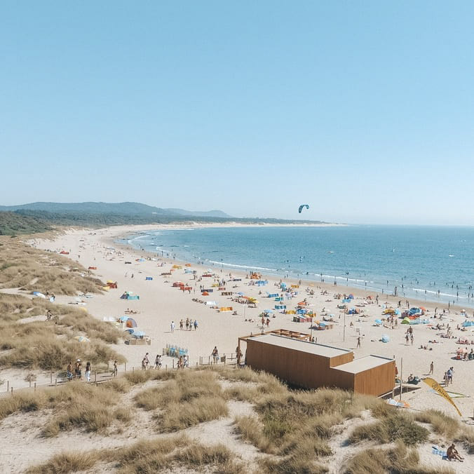 Vista aérea para o Bar de Praia do FeelViana, num dia bastante solarengo. Há banhistas dentro e fora da água e kites no ar. 
